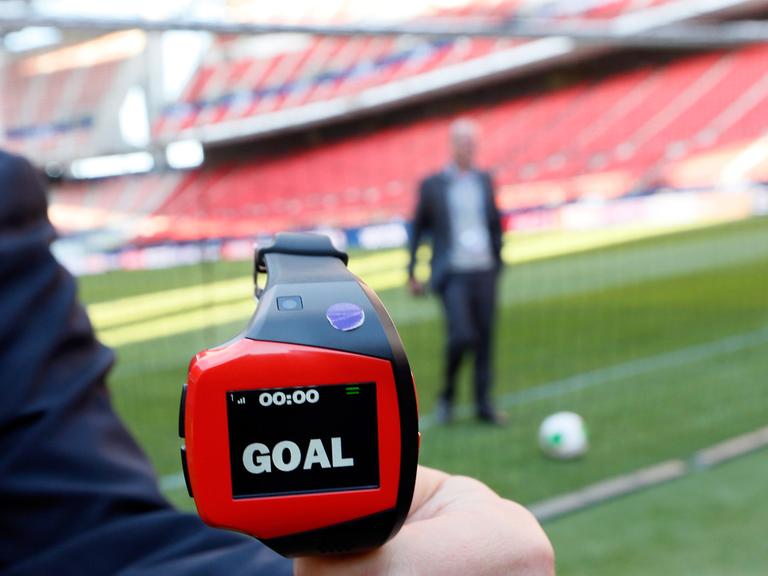 Eine Art Armbanduhr mit der Aufschrift "Goal", im Hintergrund ein Ball in einem Tor in einem Fußballstadion.