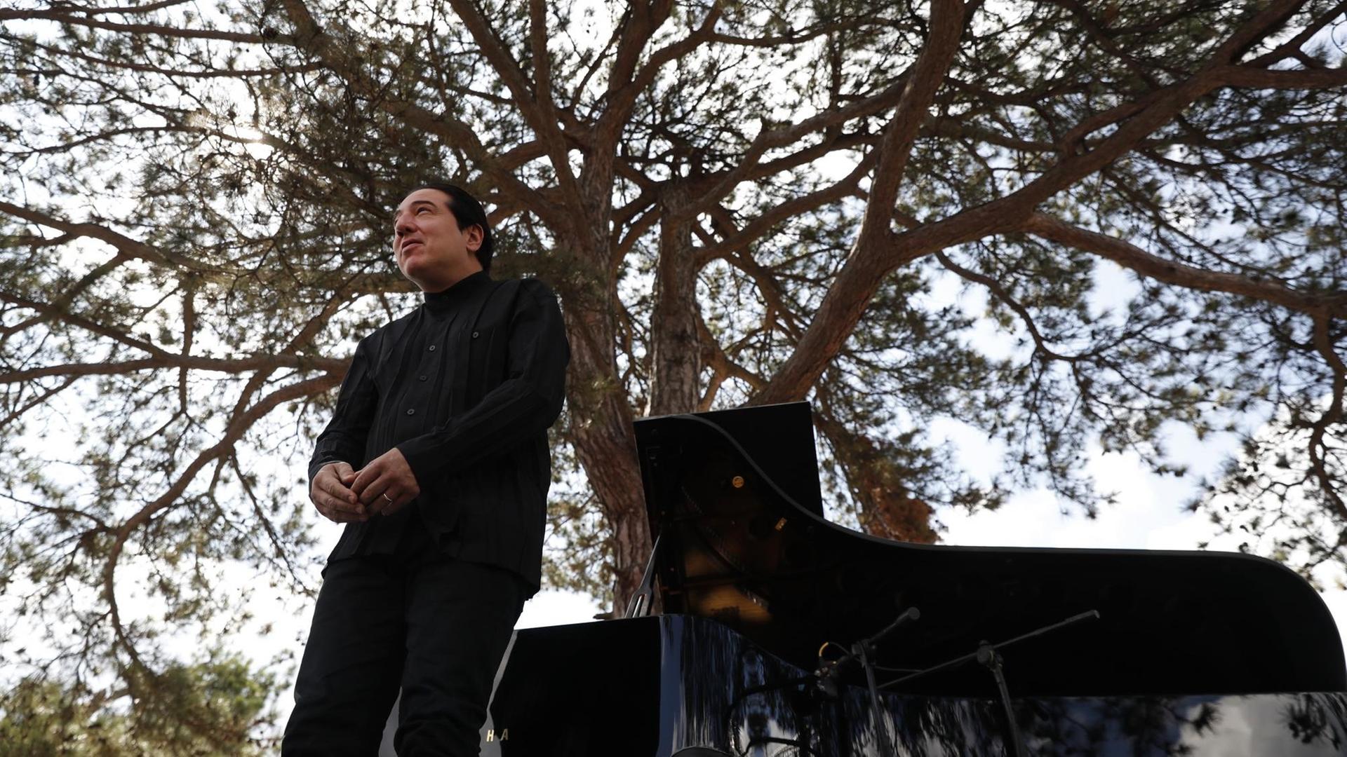Fazil Say bei einer Outdoor-Konzertveranstaltung in Kirazli, bei der er auf der Bühne vor seinem Klavier steht und sich verbeugt. Im Hintergrund steht ein großer Baum, der scheinbar aus dem Flügel erwächst.