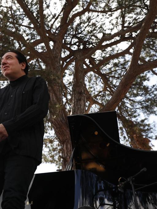 Fazil Say bei einer Outdoor-Konzertveranstaltung in Kirazli, bei der er auf der Bühne vor seinem Klavier steht und sich verbeugt. Im Hintergrund steht ein großer Baum, der scheinbar aus dem Flügel erwächst.