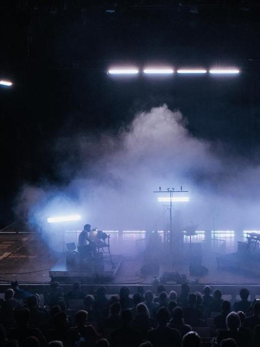 Eine in Nebel gehüllte Bühne mit drei undeutlich zu erkennenden Musikern. Im Vordergrund sieht man das Publikum, im Hintergrund Halogenleuchten.