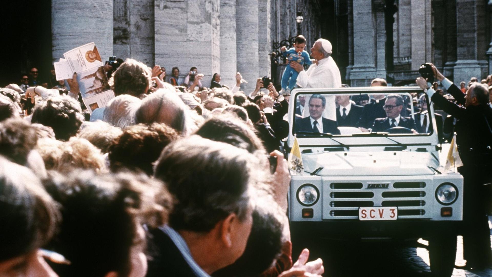 Papst Johannes Paul II. fährt am 13. Mai 1981 in einem offenen Fahrzeug an einer Menschenmenge auf dem Petersplatz in Vatikanstaat vorbei und hält ein kleines Kind auf dem Arm.