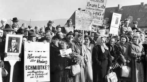 Menschen mit Suchschildern hoffen auf Nachricht von ihren Vermissten, während Bundespräsident Heuss am 18.10.1955 in Friedland Heimkehrer begrüßt.