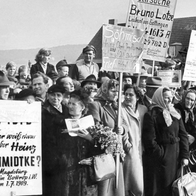 Menschen mit Suchschildern hoffen auf Nachricht von ihren Vermissten, während Bundespräsident Heuss am 18.10.1955 in Friedland Heimkehrer begrüßt. 