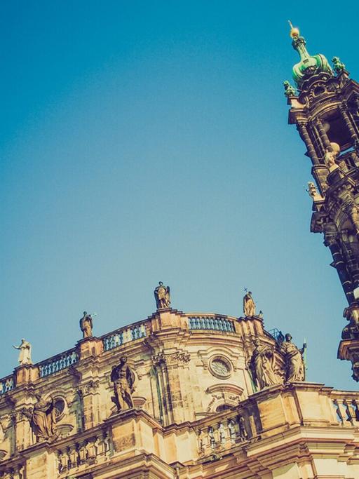 Blick von Unten zum Kirchenschiff und Turmspitze der Dresdner Hofkirche mit ihren vielen Figuren auf dem Dach und der gelb-grauen Sandsteinfassade.