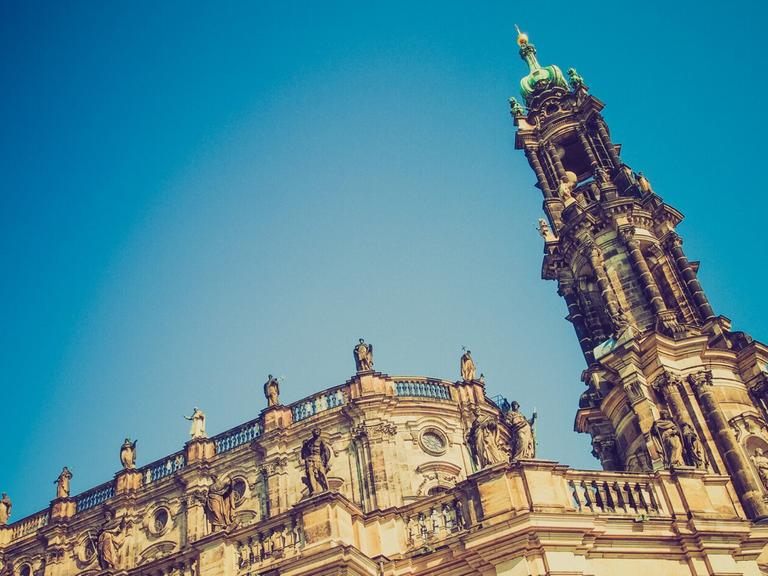 Blick von Unten zum Kirchenschiff und Turmspitze der Dresdner Hofkirche mit ihren vielen Figuren auf dem Dach und der gelb-grauen Sandsteinfassade.