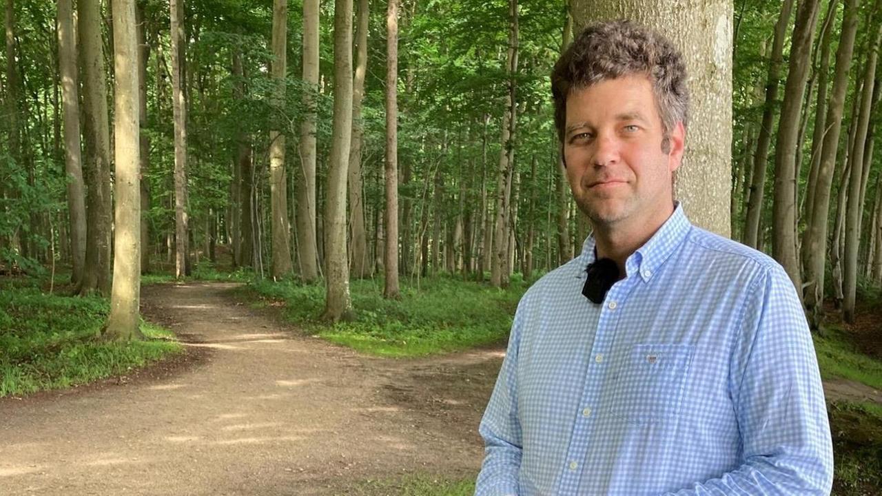 Sebastian Graf von Platen steht im Wald. Er trägt eine olivgrüne Arbeitshose und ein hellblau-weiß kariertes Hemd.