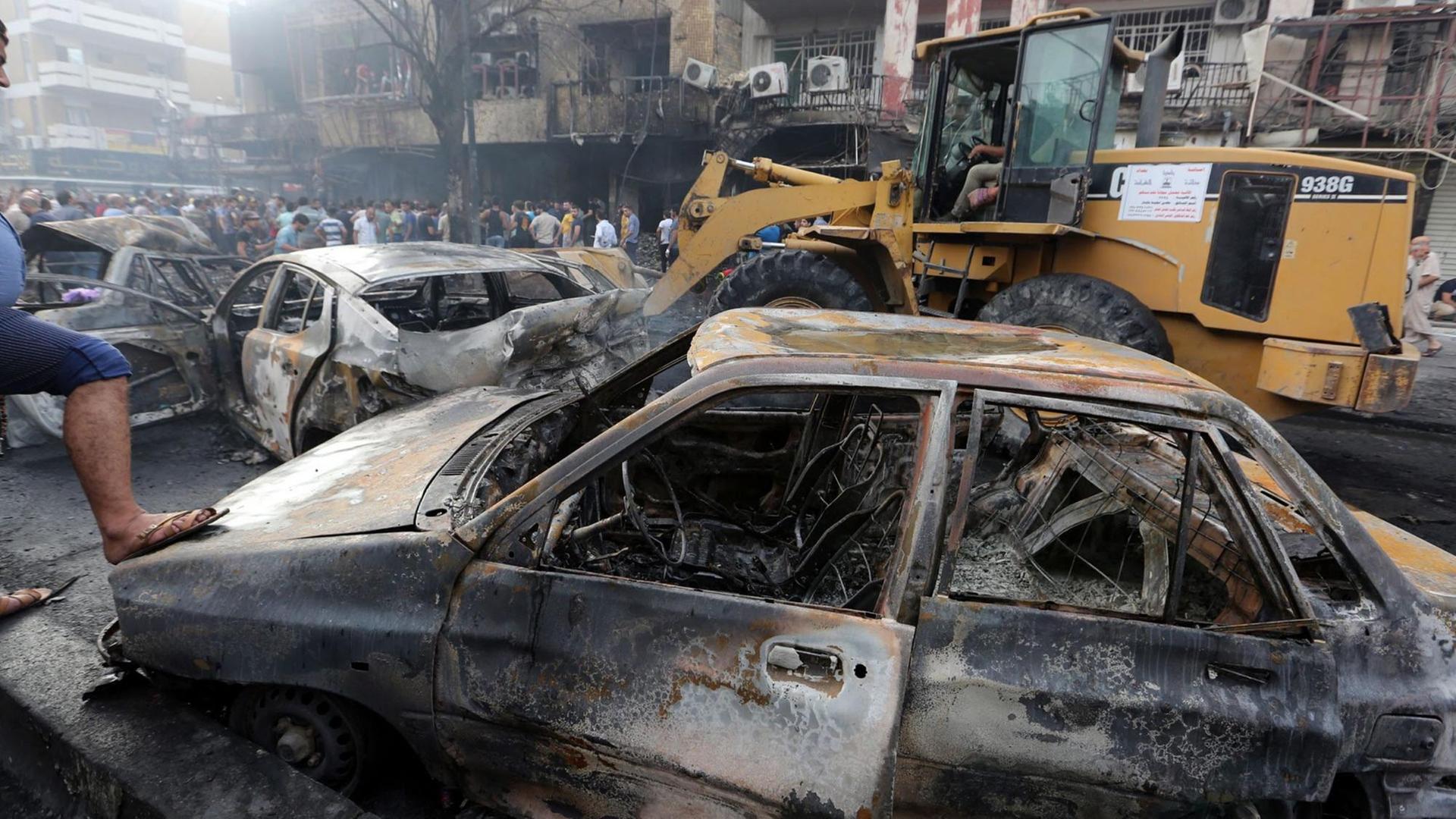 Ausgebrannte Autos vor einem beschädigten Gebäude, dazwischen Menschen