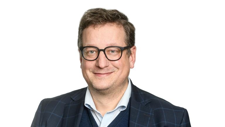 Ein bebrillter Mann in Hemd und Sakko steht vor einem weißen Hintergrund und lächelt in die Kamera. Ein bebrillter Mann in Hemd und Sakko steht vor einem weißen Hintergrund und lächelt in die Kamera.
