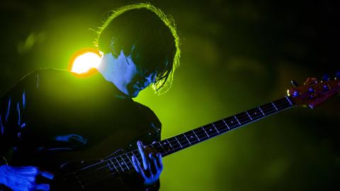 Nikolai Fraiture steht vor einem grell grünen Scheinwerfer und spielt Gitarre bei einem Auftritt von "The Strokes".