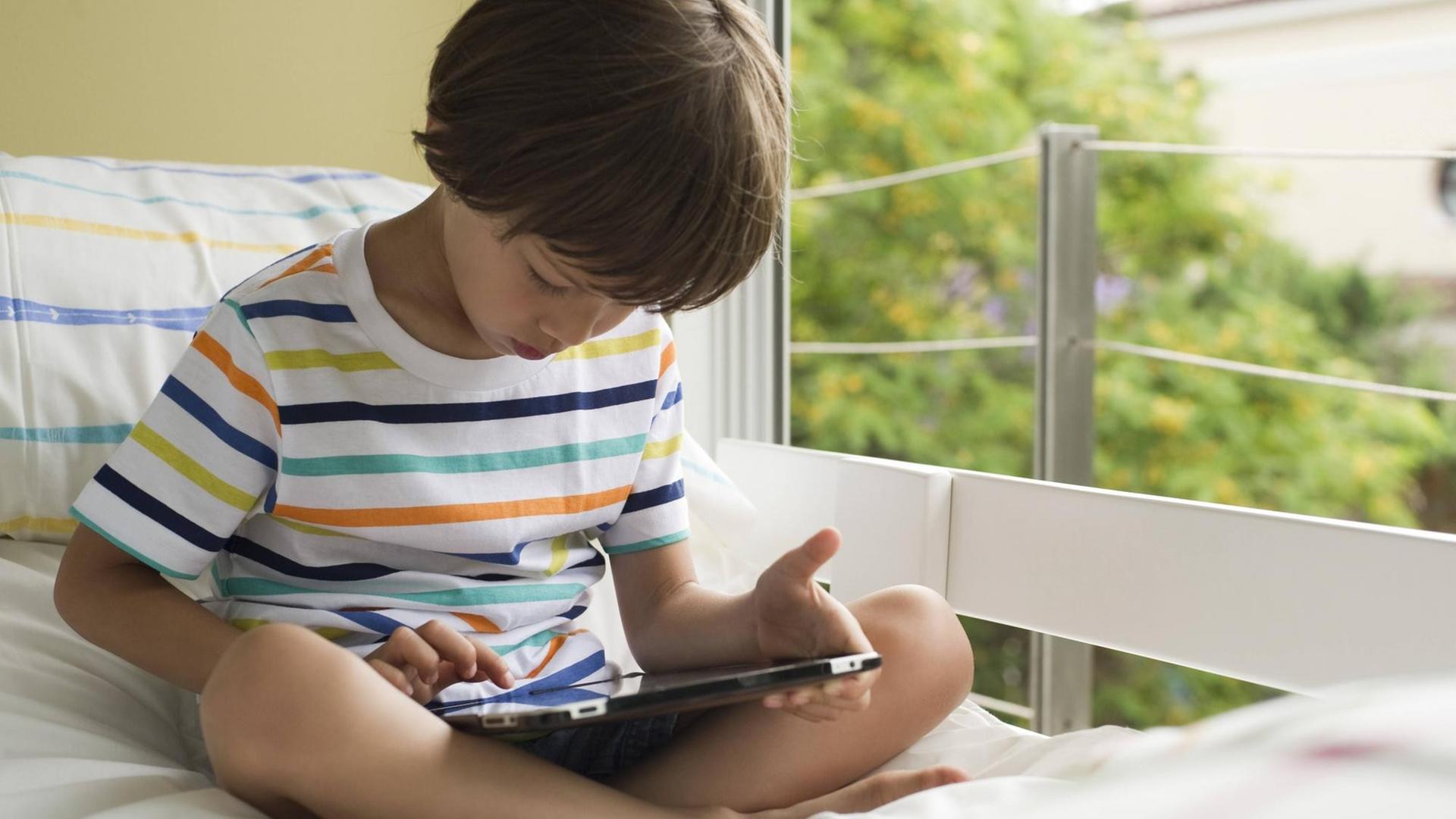 Ein Junge sitzt mit einem Tablet in der Hand auf einem Bett. Ein Junge sitzt mit einem Tablet in der Hand auf einem Bett.