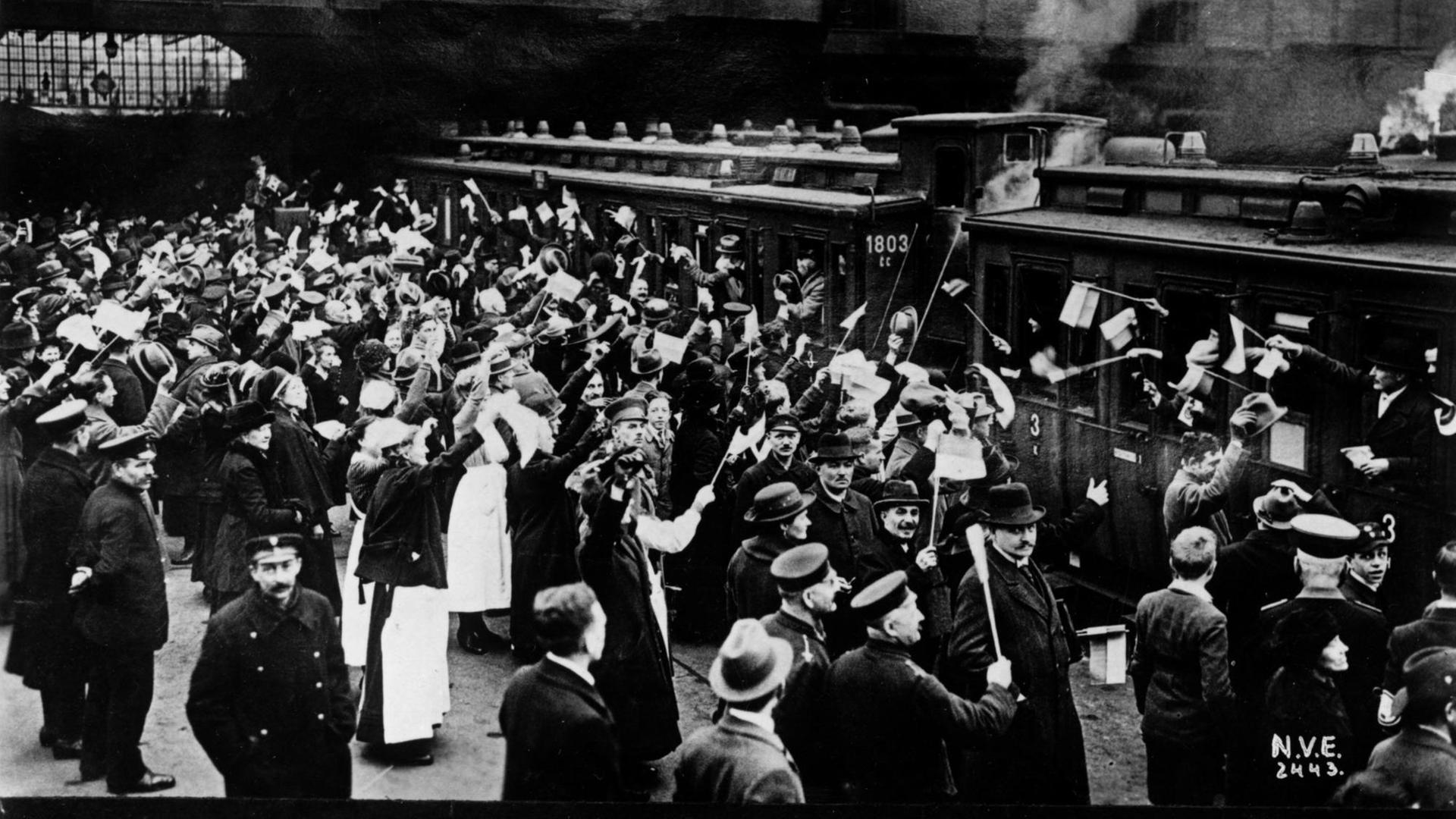 Ein Schwarzweiß-Foto zeigt eine große Menschenmenge am Gleis in einer großen Bahnhofshalle ,zum Großteil Fahnen schwenkend vor einem Zug aus zwei Waggons. Links sind zahlreiche Frauen in weißen Röcken, vermutlich einer Tracht zu sehen die dem Zug entgegen winken. Auf der rechten Bildhälfte bfinden sich zahlreiche Männer in Anzug und mit Schirmmützen Ein Sonderzug für Stimmberechtigte bei der Volksabstimmung Schlesien am 20. Maäz 1921 wartet im Schlesischen Bahnhof Berlin