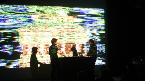 Das Bild zeigt zwei Personen am Laptop, die in einem abgedunkeltem Raum vor einer großen Leinwand stehen. Auf der Leinwand sind verzerrte bunte Streifen zu sehen.