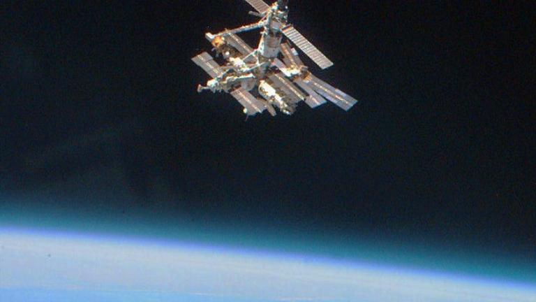 Ein Farbfoto zeigt den Blick von derUS-Raumfähre Atlantis auf die russische Raumstation Mir, einem metallenen Corpus von dem mehrere längliche Solarpaneele abgehen.Auf der oberen Bildhälfte das Schwarz des Weltalls, im scharfen Kontrast darunter das leuchtende, helle Blau der Erde