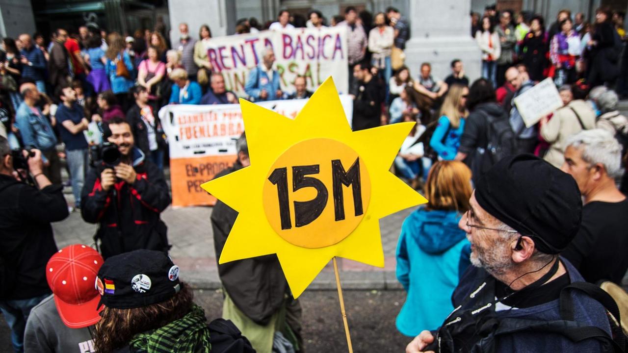 Demonstranten mit einem Schild in Form einer Sonne und mit der Aufschrift "15M".