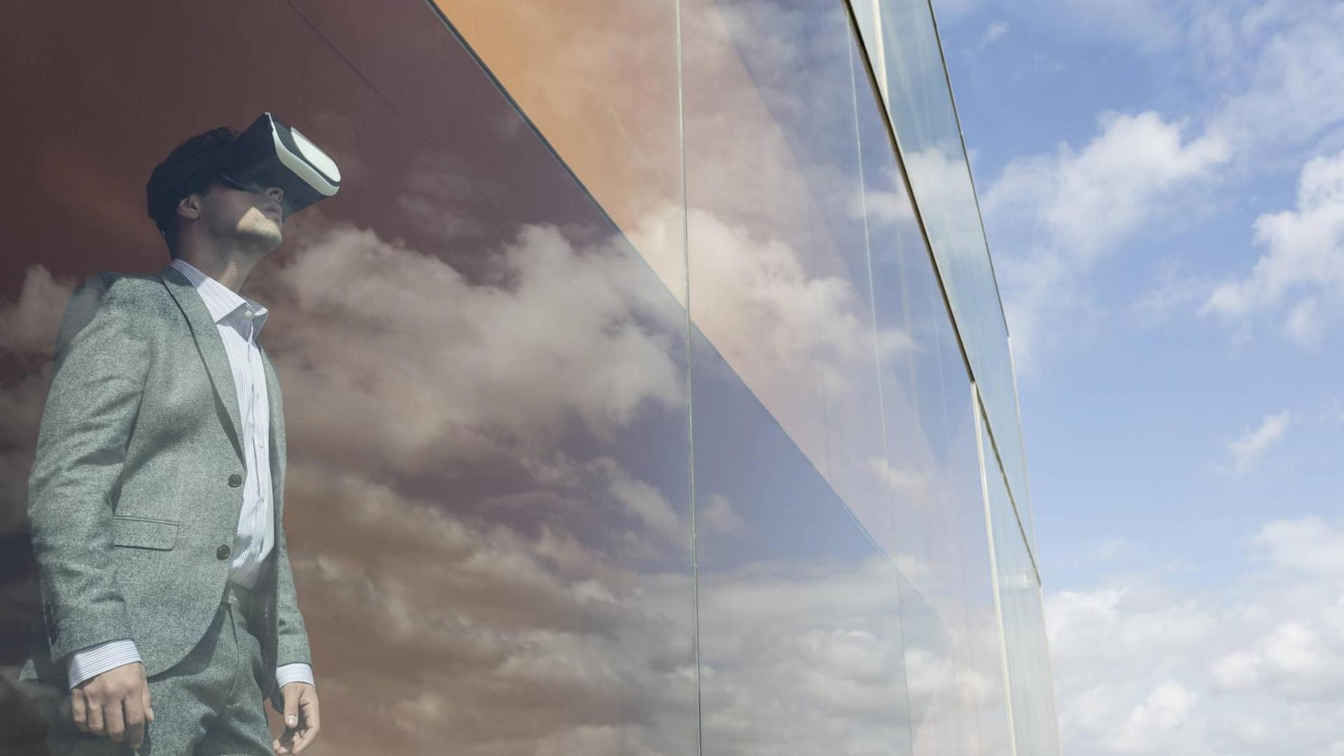 Ein Mann im Anzug und einer VR-Brille steht an einem Fenster eines Hochhauses, in dem sich der Himmel spiegelt.