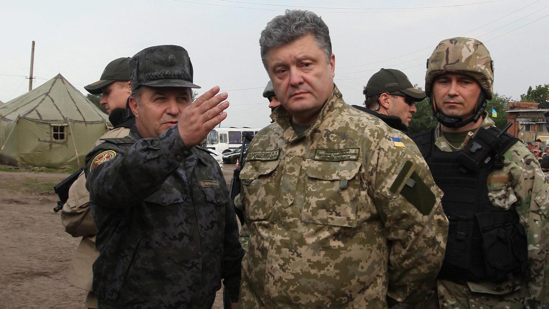 Der ukrainische Präsident Petro Poroschenko steht im Feldanzug gekleidet in einem Militärlager. Neben ihm ein Soldat, der mit ausgestrecktem Arm auf etwas außerhalb des Bildes deutet.