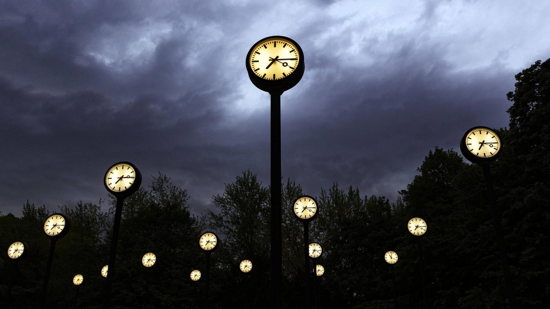 Mehrere Uhren mit beleuchtetem Zifferblatt stehen vor einem Wolkenhimmel in der Dämmerung.