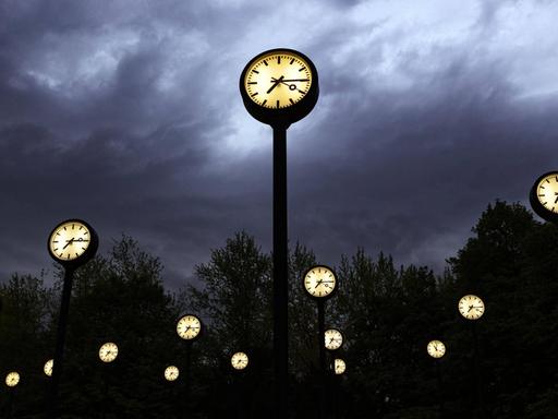 Mehrere Uhren mit beleuchtetem Zifferblatt stehen vor einem Wolkenhimmel in der Dämmerung.
