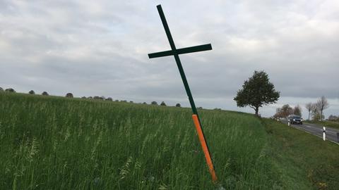 Ein grünes Holzkreuz steht auf einer Wiese neben einer Straße.