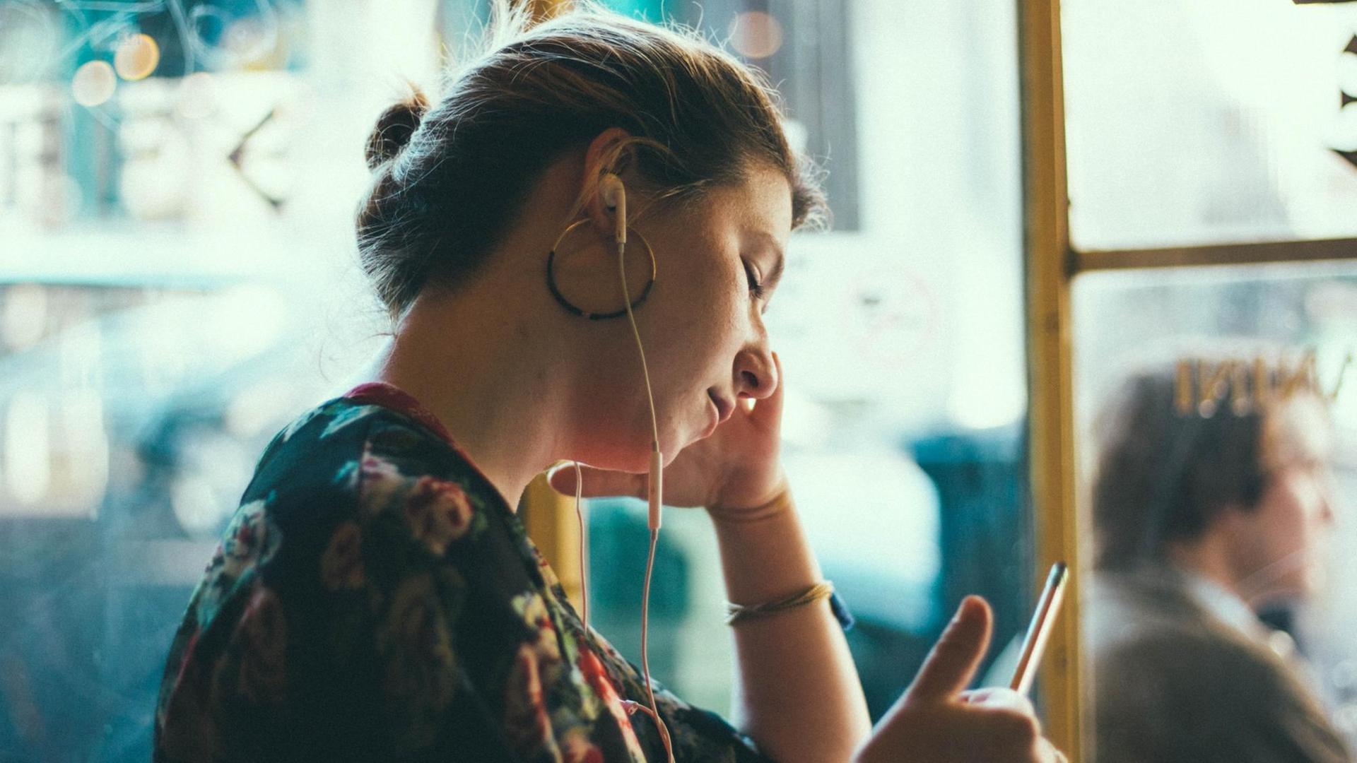 Eine Frau sitzt an einem Fenster, sie hat weiße Kopfhörer in ihren Ohren und hört eine Sendung.
