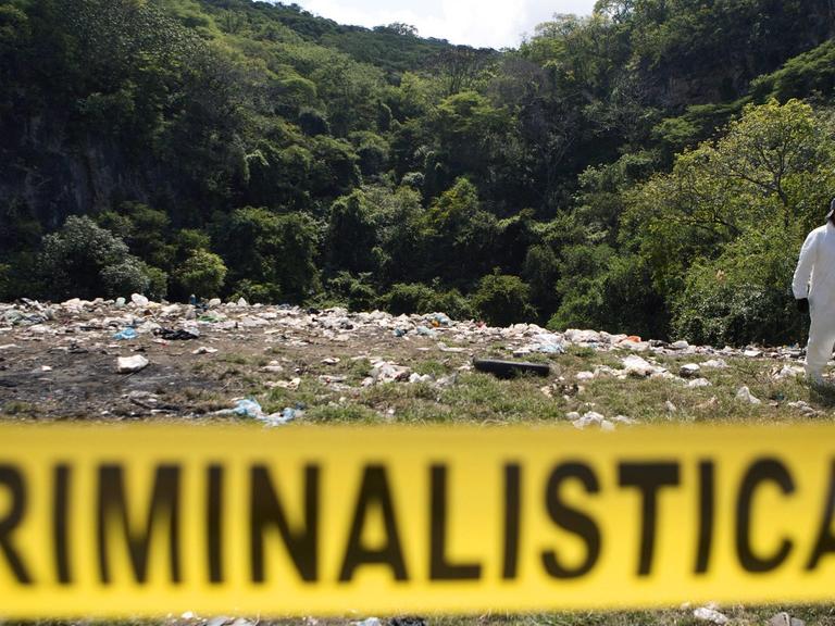 Hinter einer gelben Absperrung mit der schwarzen Aufschrift "Criminalistica" sieht man eine Müllkippe mit einem in weiße Schutzkleidung gehüllten Forensiker, im Hintergrund ein bewaldeter Hügel.
