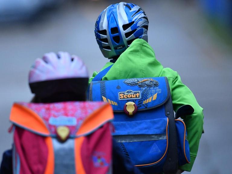 Zwei Kinder mit Schulranzen und Fahrradhelmen sind von hinten auf ihren Fahrrädern auf dem Schulweg zu sehen.