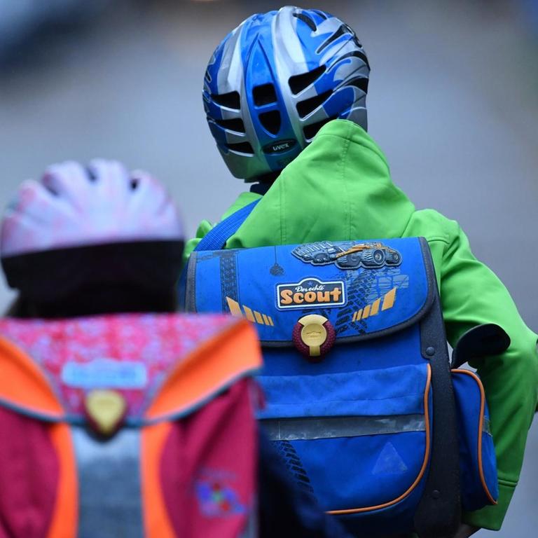 Zwei Kinder mit Schulranzen und Fahrradhelmen sind von hinten auf ihren Fahrrädern auf dem Schulweg zu sehen.