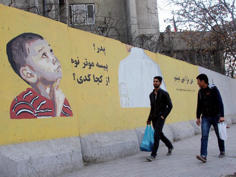 Zwei Afghanen passieren eine Mauer in Kabul mit einem Wandgemälde der Künstler und Aktivisten ArtLords: Ein kleiner Junge schaut darauf zu seinem Vater auf, auf Dari steht daneben: Vater, von wem hast du das Geld für dein Auto bekommen? (aufgenommen 2016)