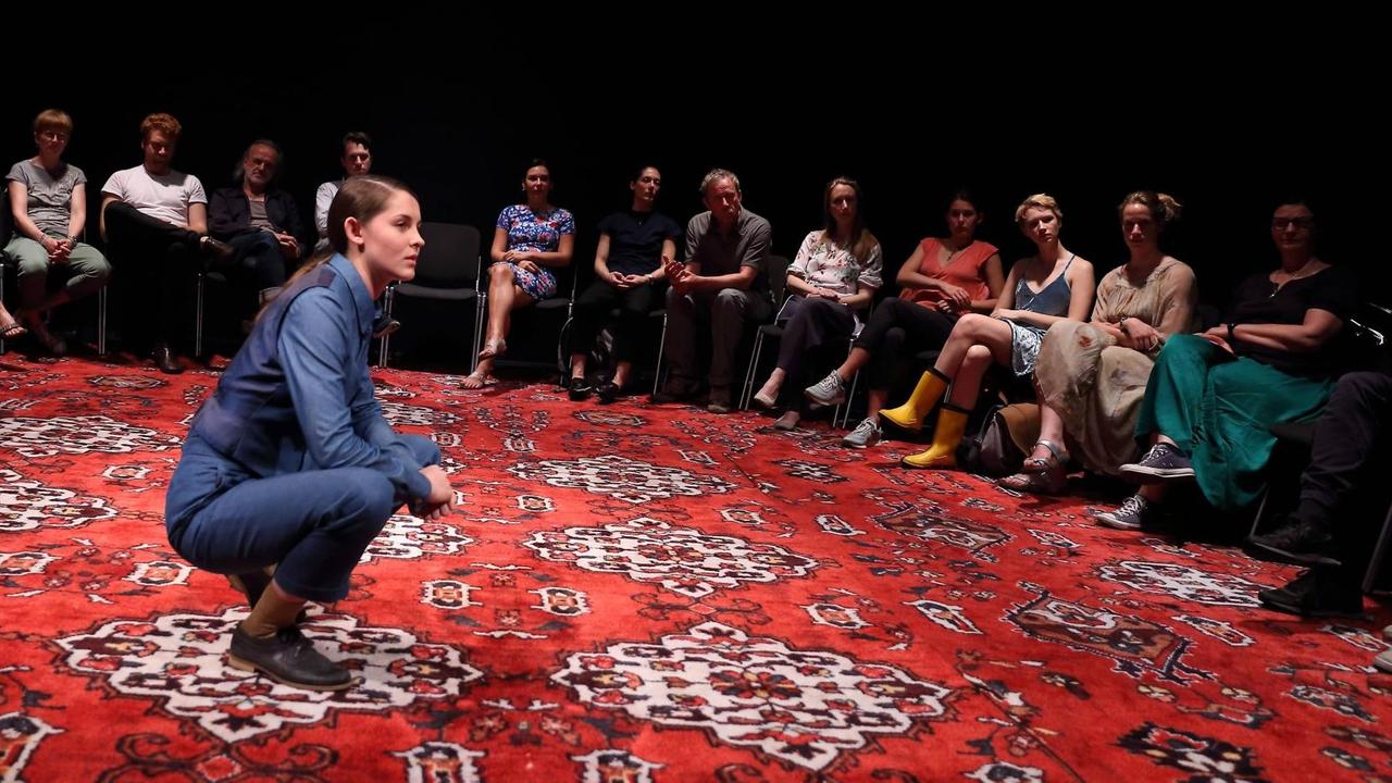 Lorena Handschin (als Kim) kniet während der Fotoprobe zu "zu unseren füßen, das gold, aus dem boden verschwunden" von Svealena Kutschke in der Regie von Andras Dömötör auf einem Teppich, um sie herum ein Stuhlkreis, in dem Frauen und Männer sitzen, aufgenommen am 6. Juni 2019 in der Box des Deutschen Theaters Berlin