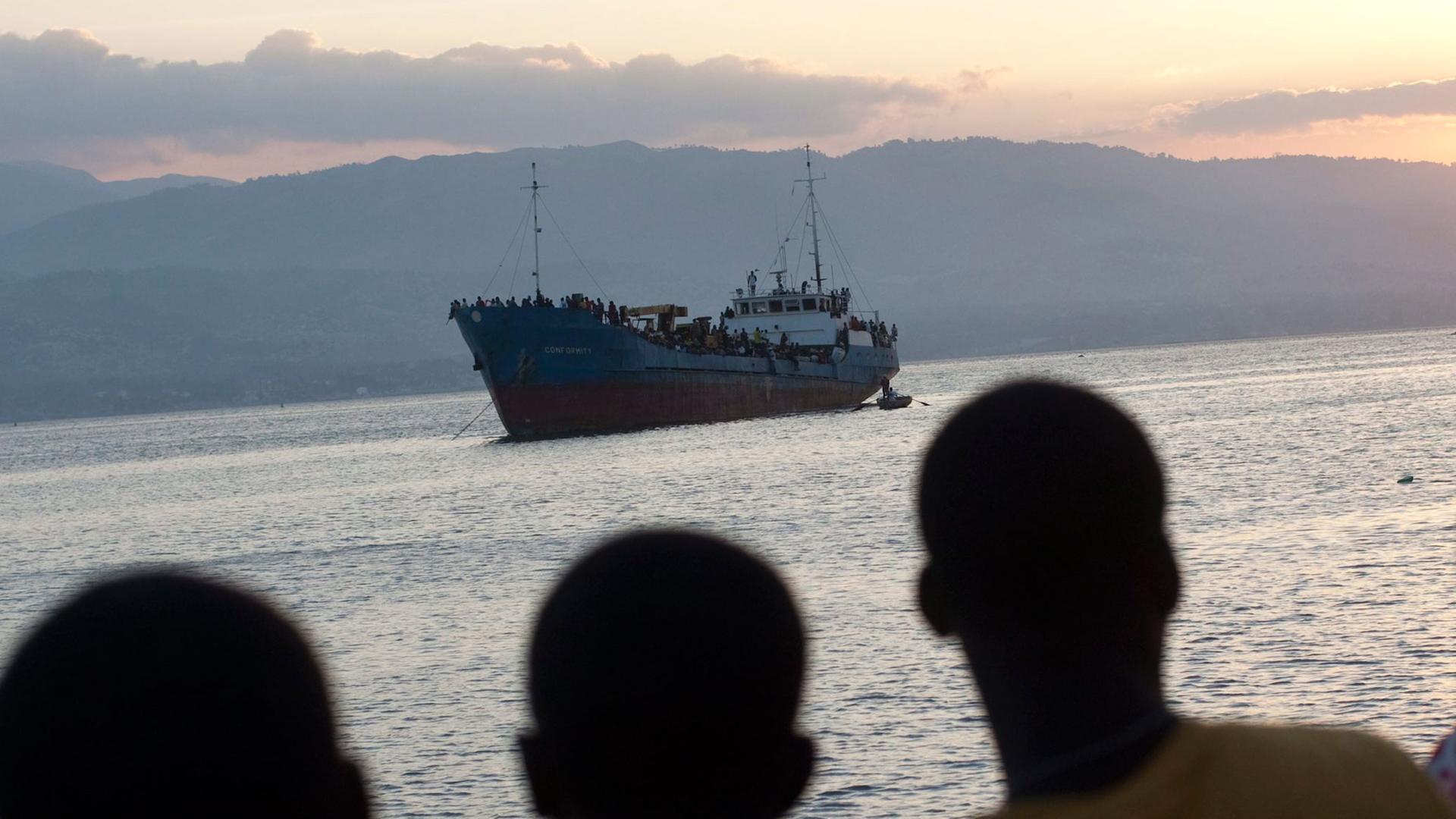 Junge Männer auf Haiti betrachten ein Schiff, auf dem sich viele Flüchtlinge befinden.