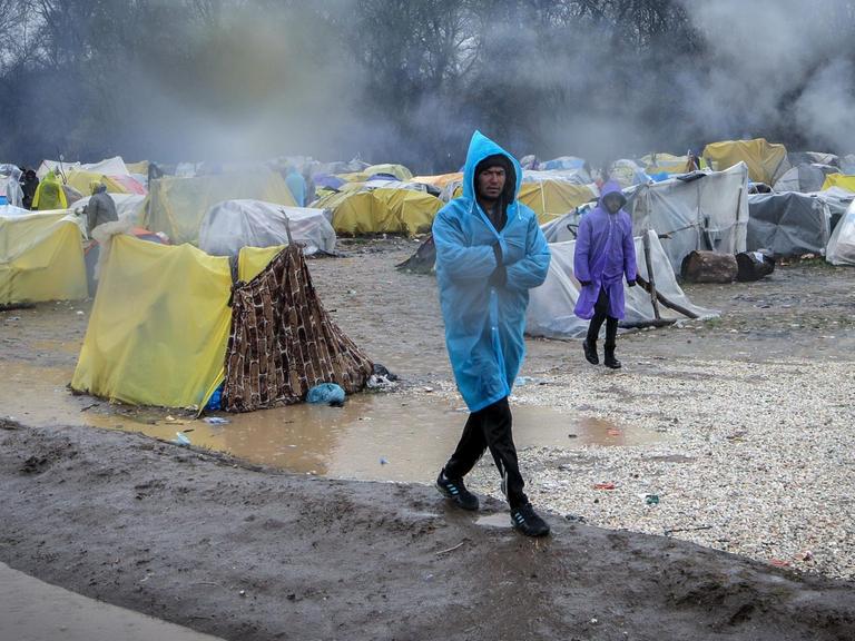 Zwei Männer im Regenponcho gehen durch ein notdürftiges Zeltlager von Flüchtlingen.