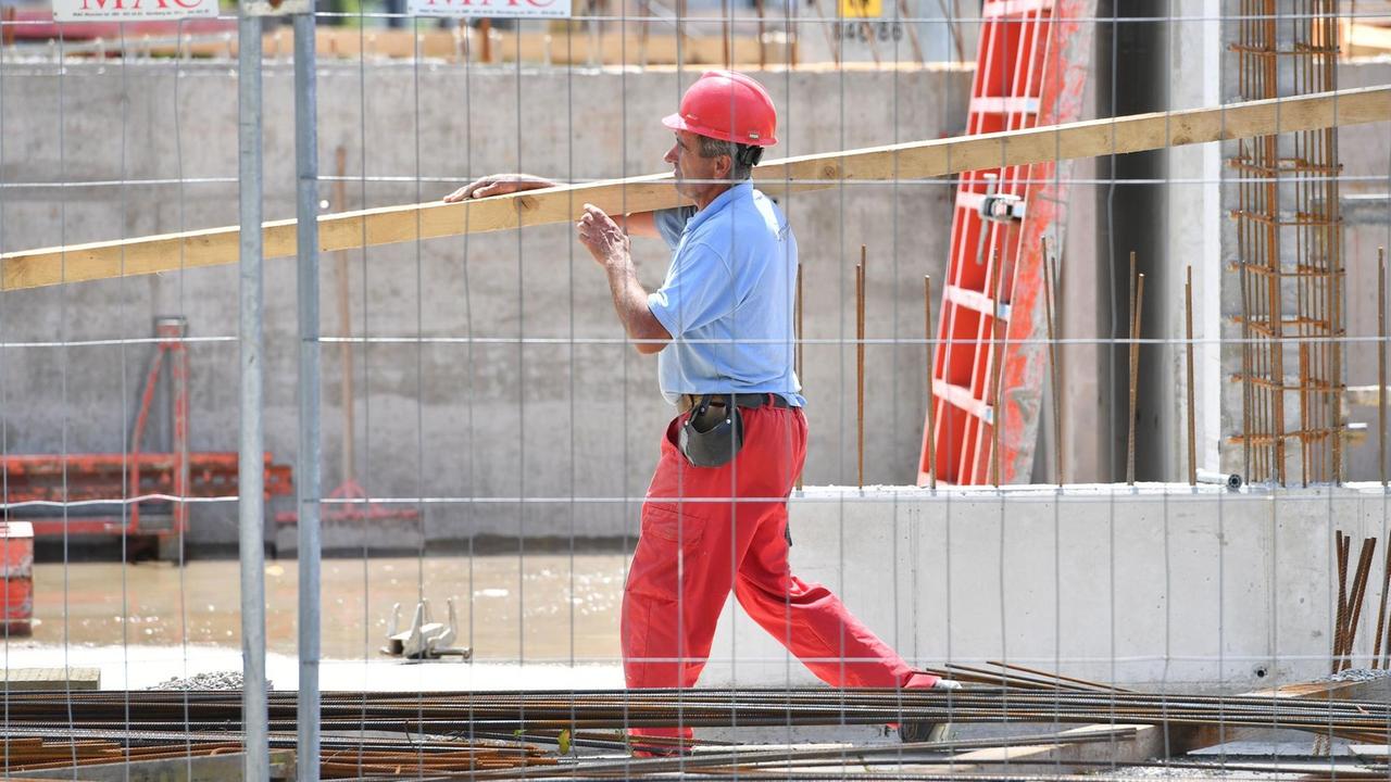 Ein Arbeiter trägt auf einer Großbaustelle eine Holzlatte. Er hat einen Helm auf und trägt ein Shirt.