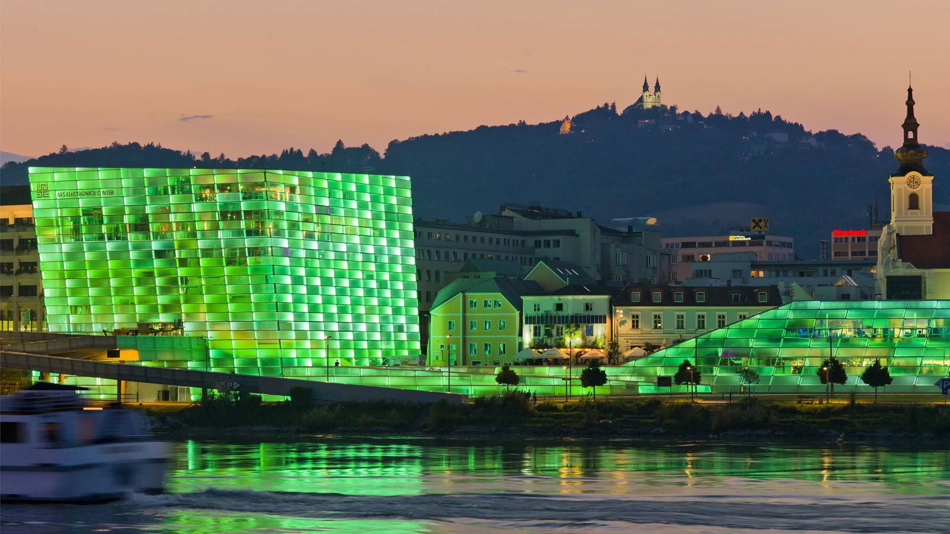 Blick auf das Ars Electronica Center in Linz mit seiner in grünes Licht getauchten Fassade. Das Gebäude spiegelt sich im Wasser, im Hintergrund ist ein Berg zu sehen.