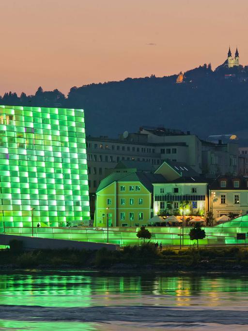 Blick auf das Ars Electronica Center in Linz mit seiner in grünes Licht getauchten Fassade. Das Gebäude spiegelt sich im Wasser, im Hintergrund ist ein Berg zu sehen.