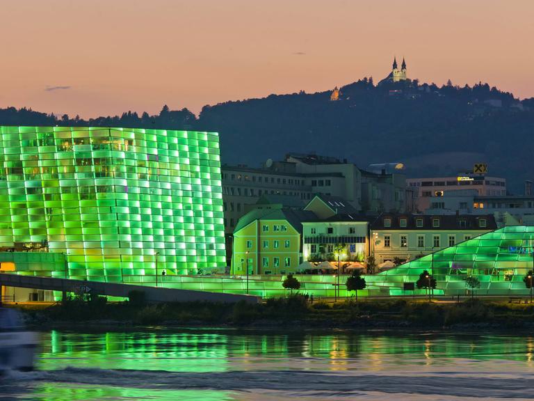 Blick auf das Ars Electronica Center in Linz mit seiner in grünes Licht getauchten Fassade. Das Gebäude spiegelt sich im Wasser, im Hintergrund ist ein Berg zu sehen.