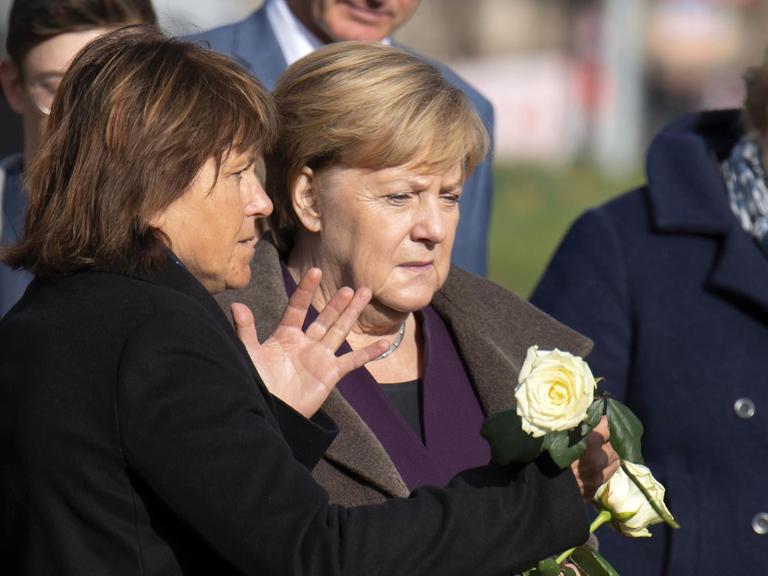 Die drei stehen nebeneinander, Merkel und Findeiß halten weiße Rosen in den Händen. Findeiß sagte etwas zu Merkel und gestikuliert mit der anderen Hand. Dahinter weitere Personen.