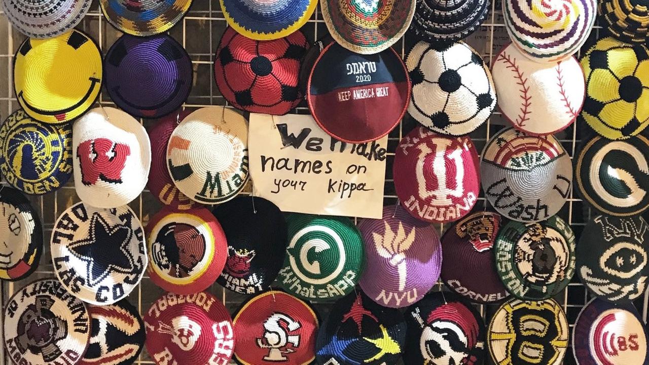 Kippas mit bunten Mustern vor einem Laden in Jerusalem. Am Ständer hängt ein Schild mit der Aufschrift: "We make names on your Kippa."