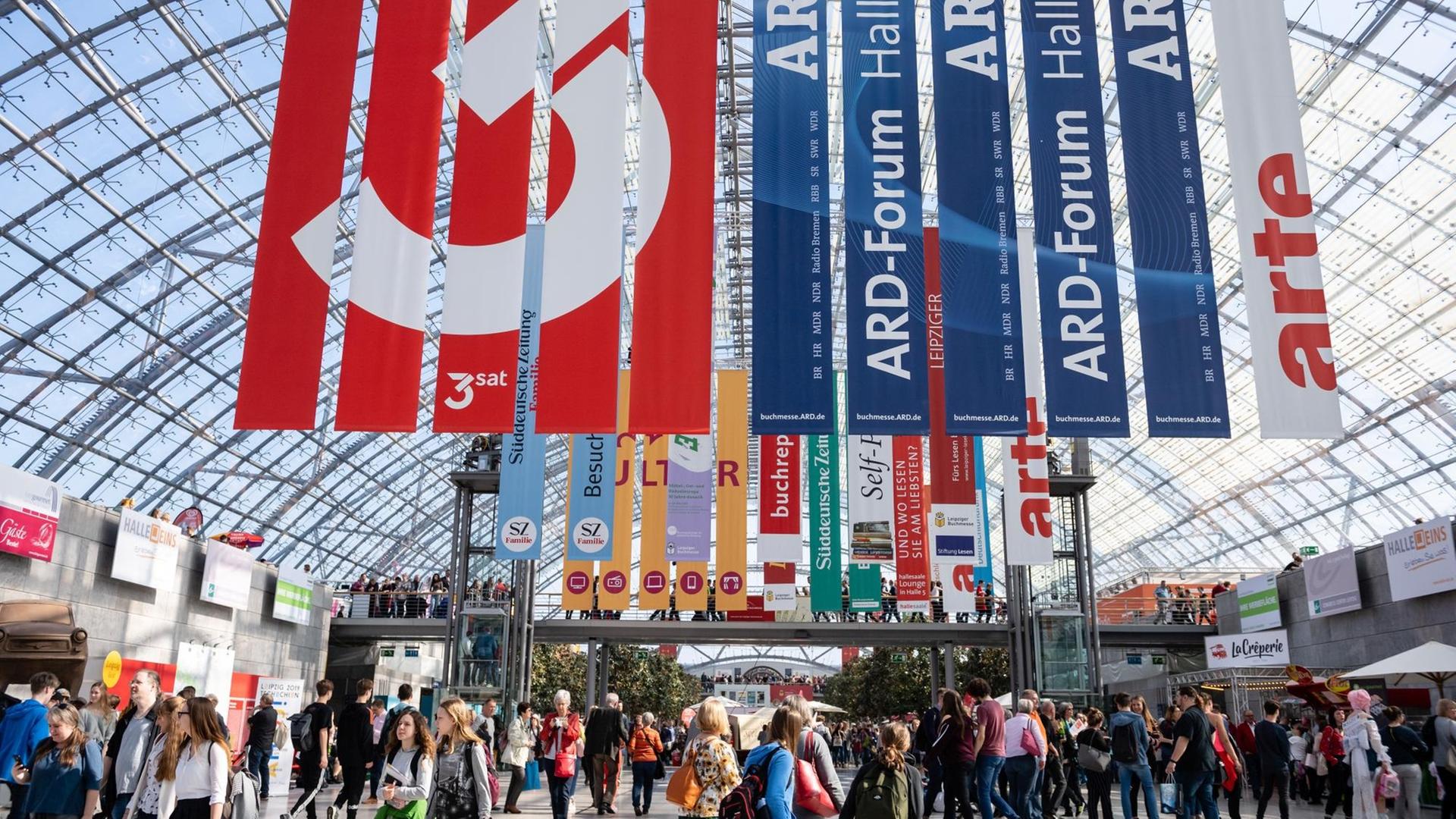 Menschen tummeln sich in einer Halle mit rundem Glasdach unter unzähligen Bannern von Verlagen und öffentlich-rechtlichen Fernsehsendern wie ARD, 3Sat oder arte. Menschen tummeln sich in einer Halle mit rundem Glasdach unter unzähligen Bannern von Verlagen und öffentlich-rechtlichen Fernsehsendern wie ARD, 3Sat oder arte.