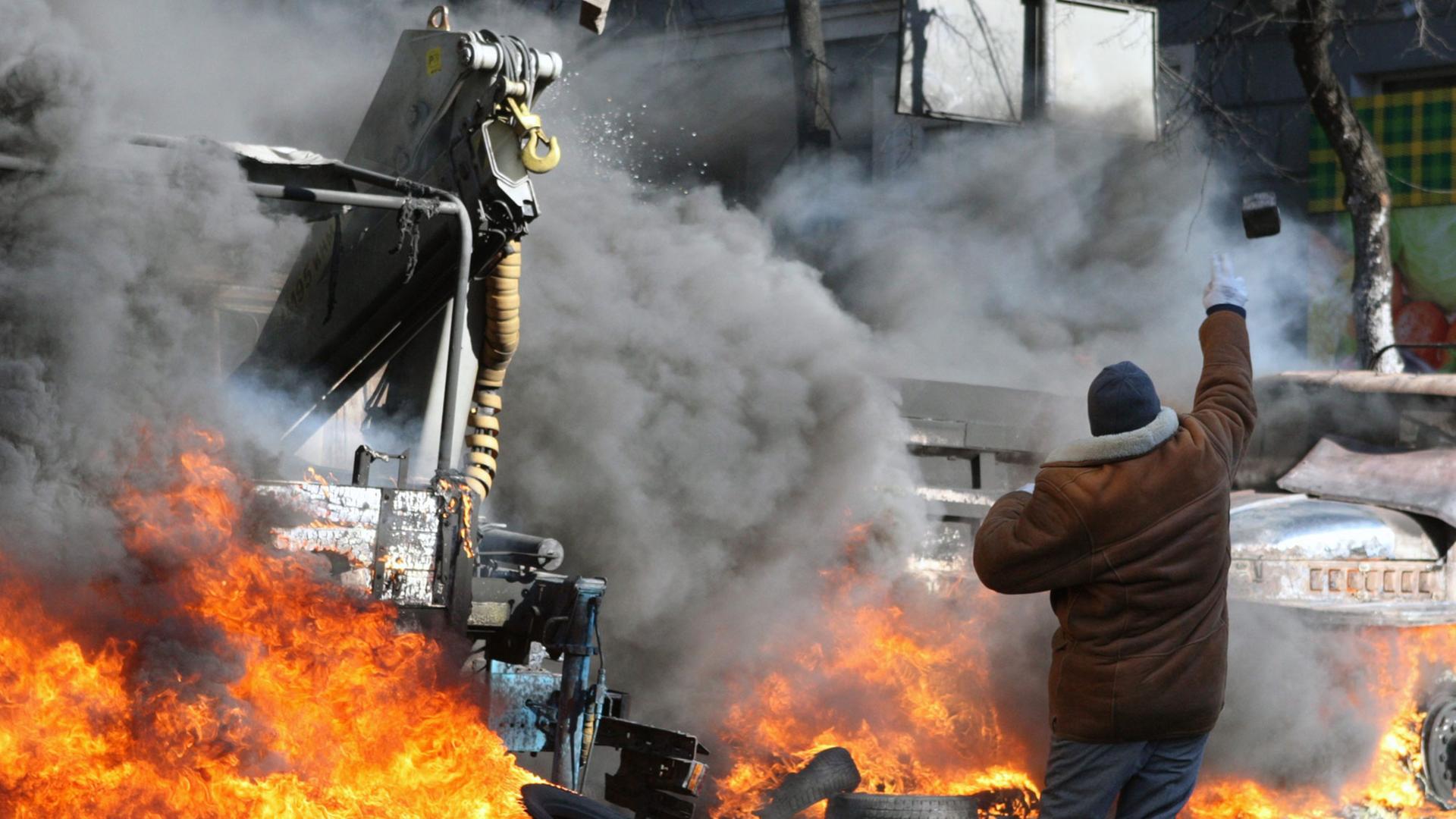 Ein Demonstrant wirft einen Pflasterstein, im Hintergrund brennt ein Auto.