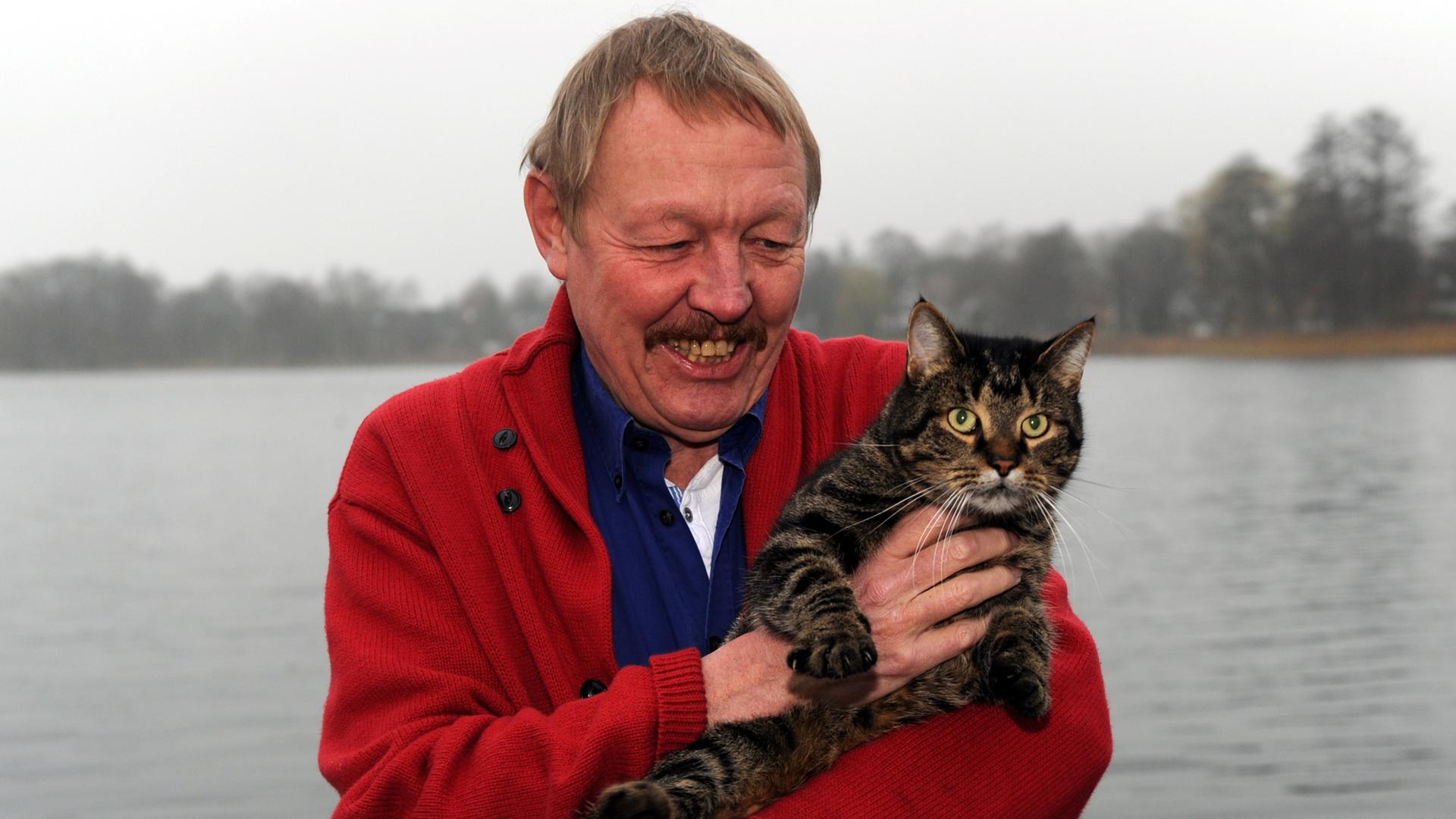 Der Gründer des Eichborn Verlags, Vito von Eichborn, steht mit seinem Kater an einem See hinter seinem Haus in Schleswig-Holstein.