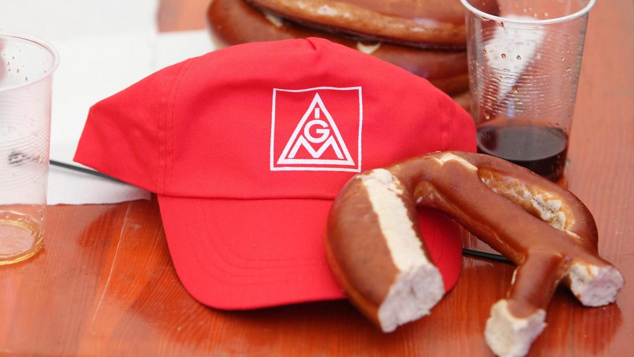 Eine rote Kappe mit IG Metall-Logo und eine angebissene Brezel liegen auf einem Tisch.