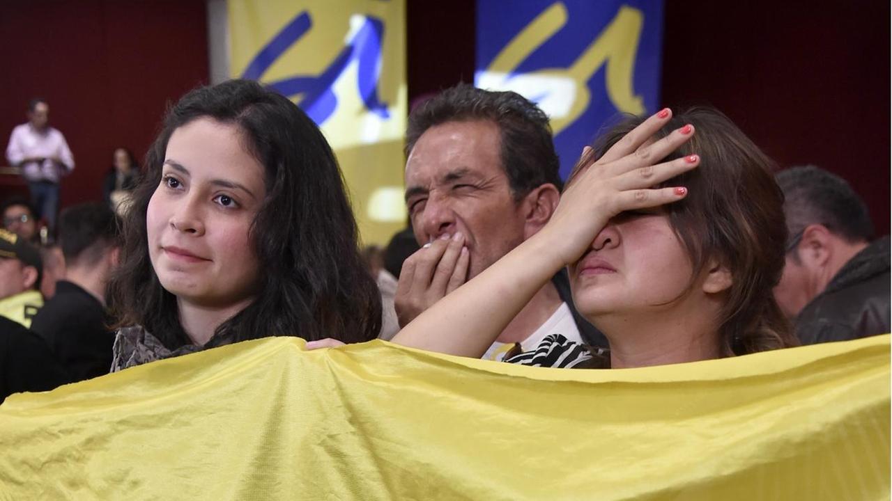 Zwei Frauen und ein Mann stehen mit enttäuschten Gesichtern hinter einer gelben Flagge. Der Mann und eine Frau schlagen die Hände vors Gesicht. 