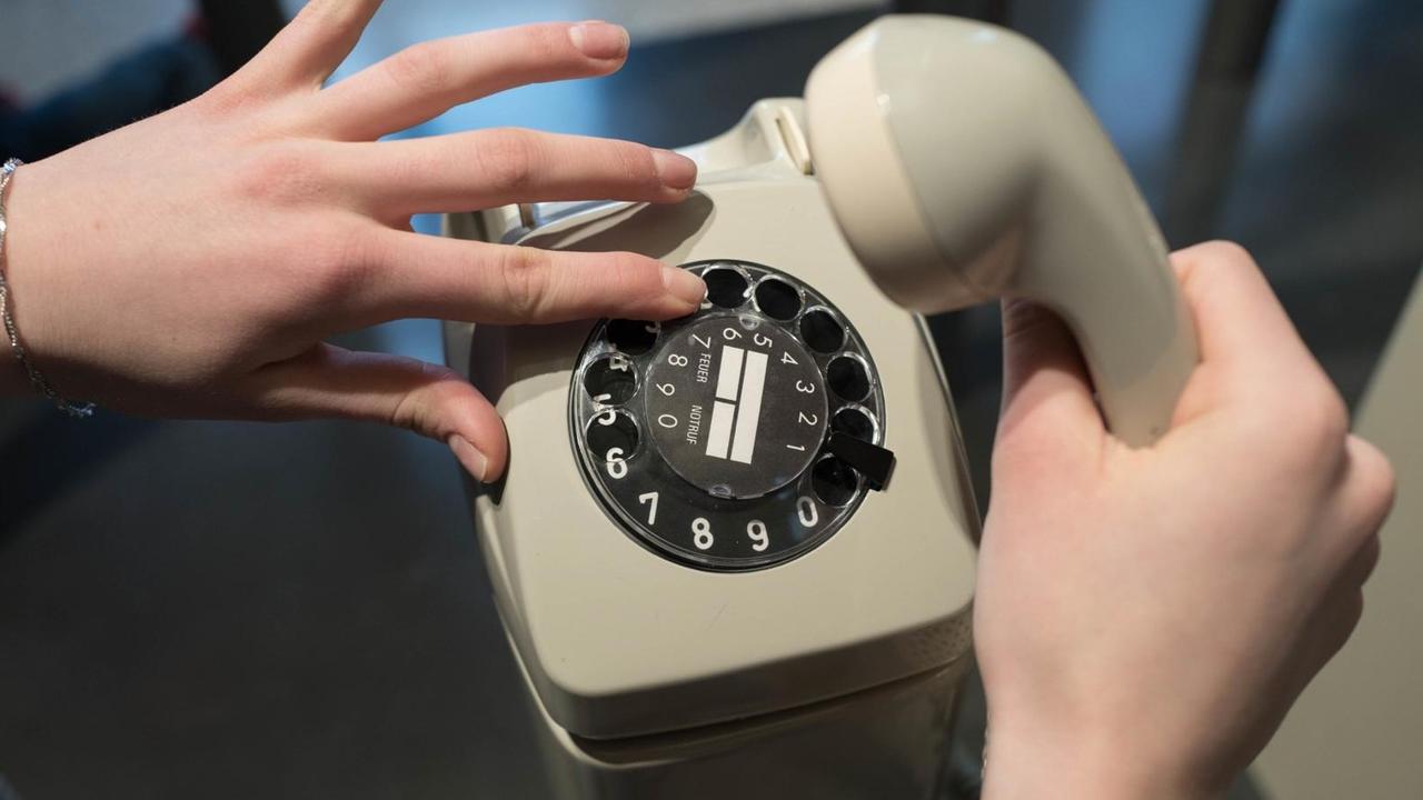 Eine linke Hand betätigte an einem grauen Telefon älteren Models die Wählscheide, die rechte hält den Hörer.