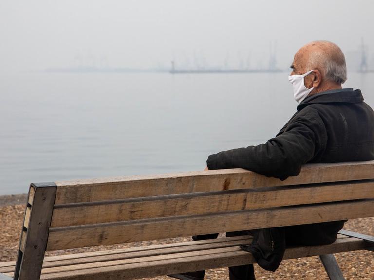 Mann mit Schutzmaske sitzt auf einer Bank an der Uferpromenade.