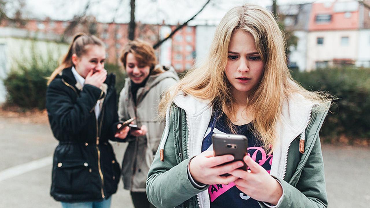 Ein Mädchen steht im Hof einer Schule und schaut auf ihr Smartphone, während im Hintergrund zwei andere Mädchen lachen.