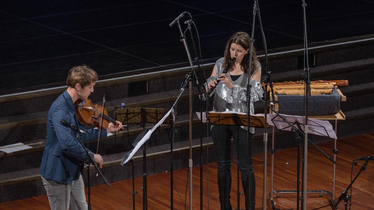 Mann mit Bratsche und Frau mit Blockflöte musizieren vor Notenständern und Mikrofonen