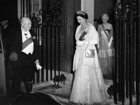 Der britische Premierminister Winston Churchill hält die Autotür für Königin Elizabeth II. auf. Seine Frau Clementine Churchill steht im Eingang von Downing Street No 10.