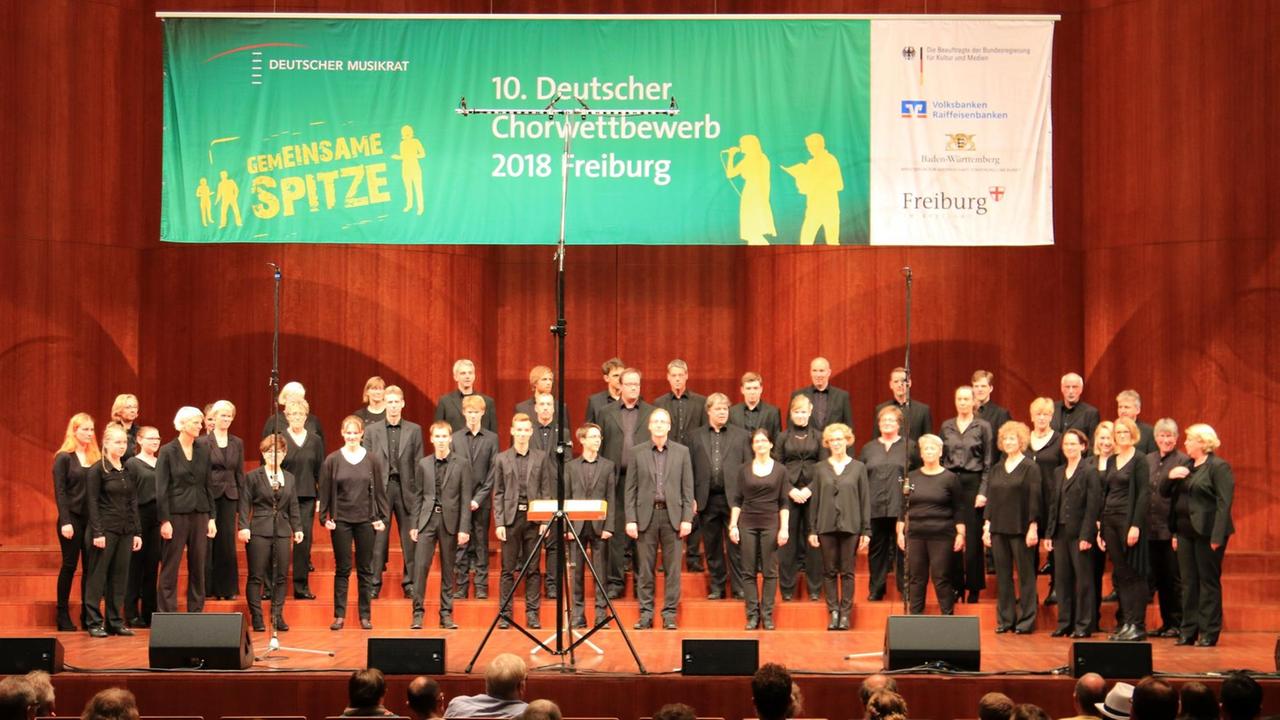 Der Chor steht beim auf der Bühne. Darüber ein großes Banner vom Chorwettbewerb in Freiburg.