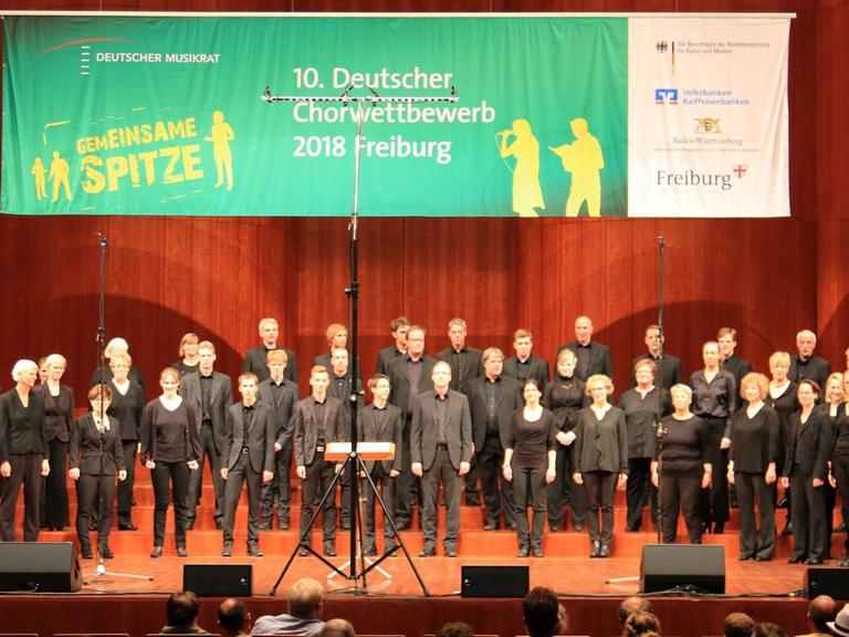 Der Chor steht beim auf der Bühne. Darüber ein großes Banner vom Chorwettbewerb in Freiburg.