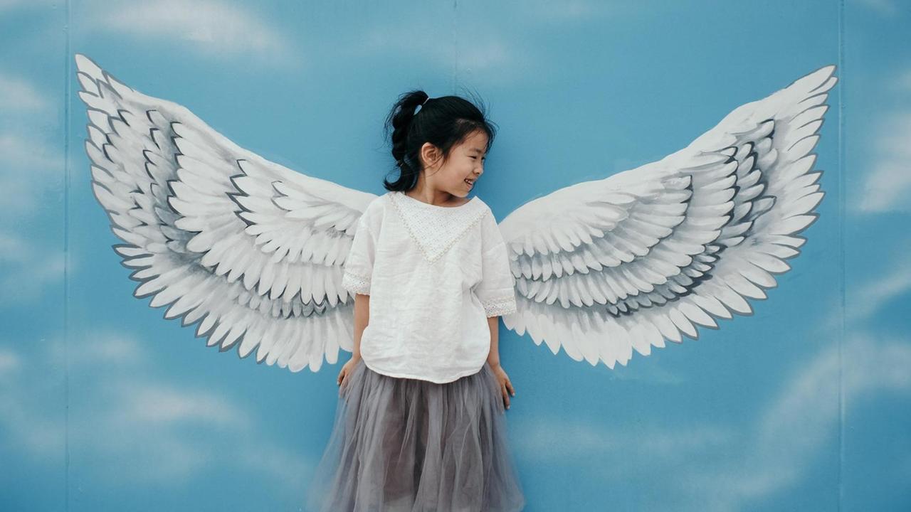 Ein kleines Mädchen steht lächelnd vor einer bemalten Wand: einem blauen Himmel, davor weisse Engelsflügel. Die Flügel sehen aus als wenn sie dem Mädchen angewachsen sind.